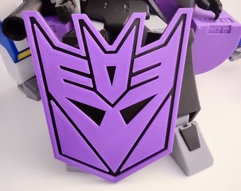 May include: Purple plastic shield with a black outline of the Decepticon symbol. The shield is held by a gray robot toy.