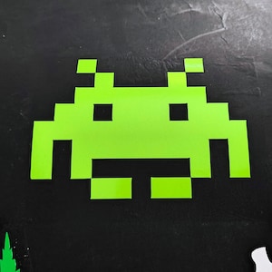 May include: A bright green pixelated alien graphic, reminiscent of classic arcade games, is centered on a black background. A white skull graphic is in the lower right corner. The image has a retro video game theme.