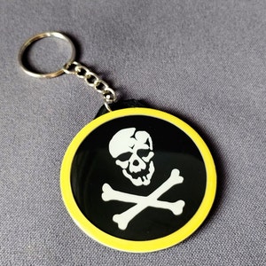 May include: A black and white skull and crossbones design on a round keychain with a yellow border. The keychain is attached to a silver chain and ring.