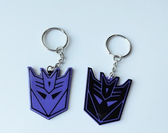 May include: Two keychains featuring the Transformers logo. One keychain is purple with a black logo, and the other is black with a purple logo.