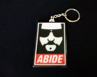 May include: Black and white image of a man's face with sunglasses and a beard on a black rectangle with the word "ABIDE" in red letters on a white background. The rectangle is attached to a silver key chain.