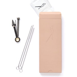 Puede incluir: Un juego de accesorios para fumar en una caja de color marrón claro. El juego incluye una herramienta de metal para pipa, tres cepillos de limpieza y un pequeño paquete. La caja tiene un logotipo con una figura estilizada.