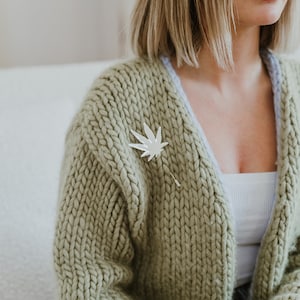 May include: A woman wearing a light green knitted cardigan with a white leaf pin. The cardigan has a light blue lining.