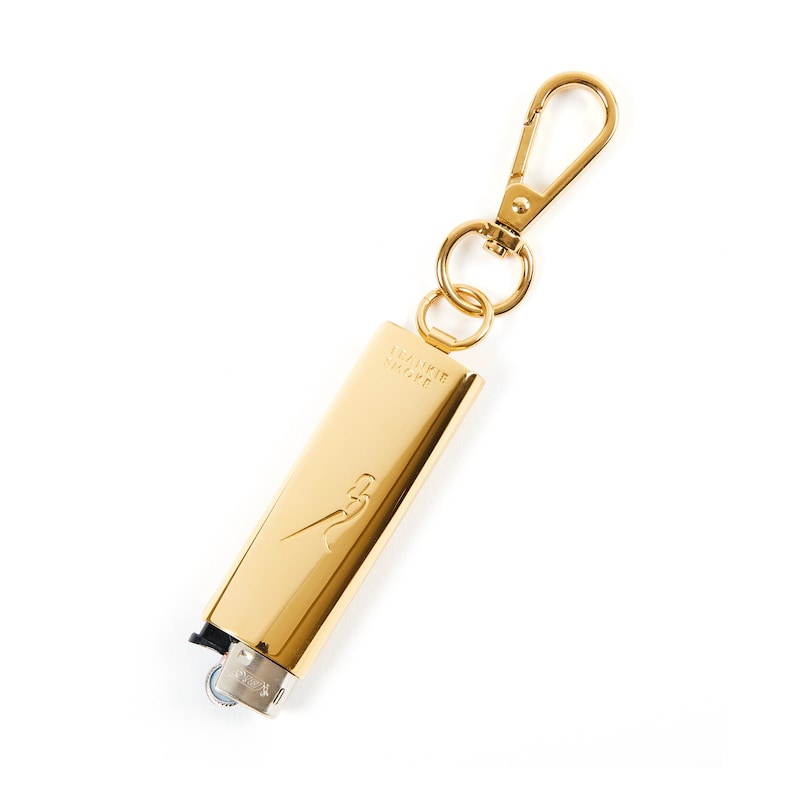 May include: A gold-coloured lighter keychain with the text "Frankie Shop" engraved on the side. The lighter has a small, silver-coloured wheel on the side.