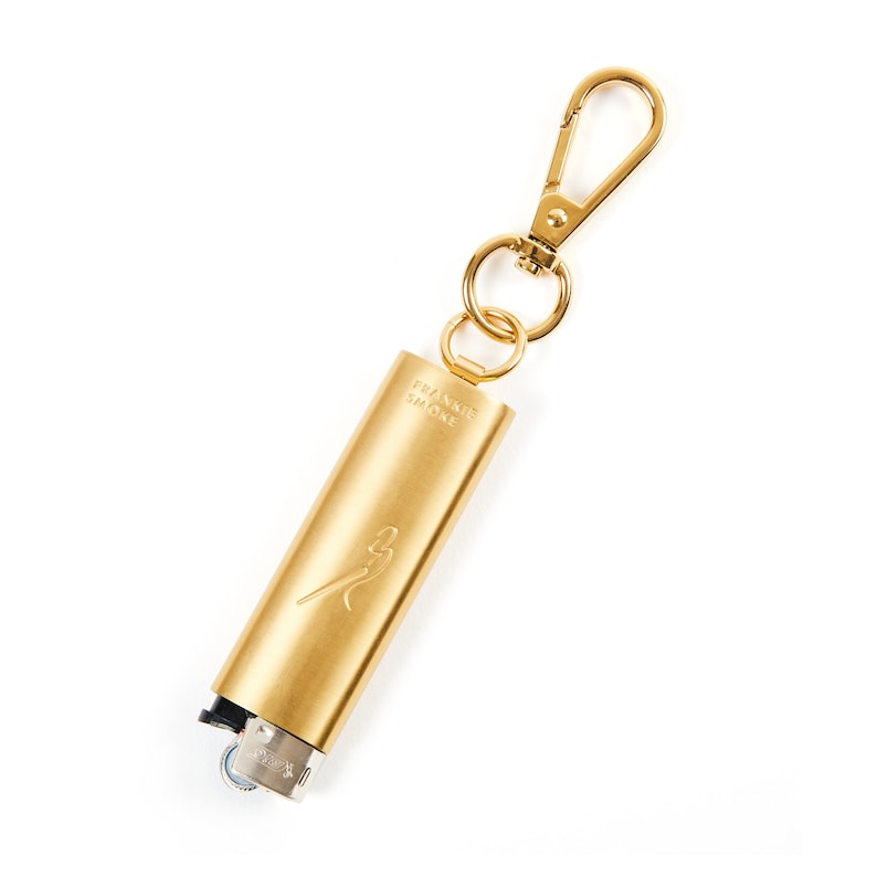 May include: A gold-coloured lighter keychain with a Frankie Shop logo and a silver lighter. The keychain has a gold-coloured metal clip.