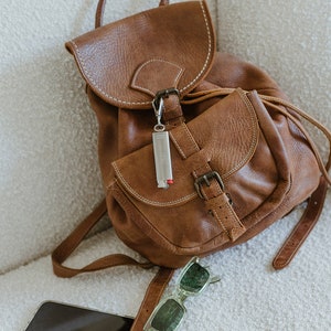 May include: A brown leather backpack with a silver keychain and a pair of green sunglasses. A black smartphone is lying on the white surface.