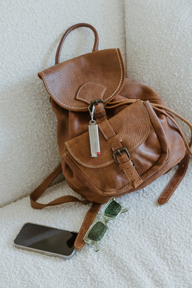 May include: A brown leather backpack with a silver keychain and a pair of green sunglasses. A black smartphone is lying on the white surface.