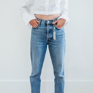 May include: A woman wearing a white cropped button-down shirt with long sleeves, blue denim jeans, and red shoes. She is standing with her hands in her pockets.