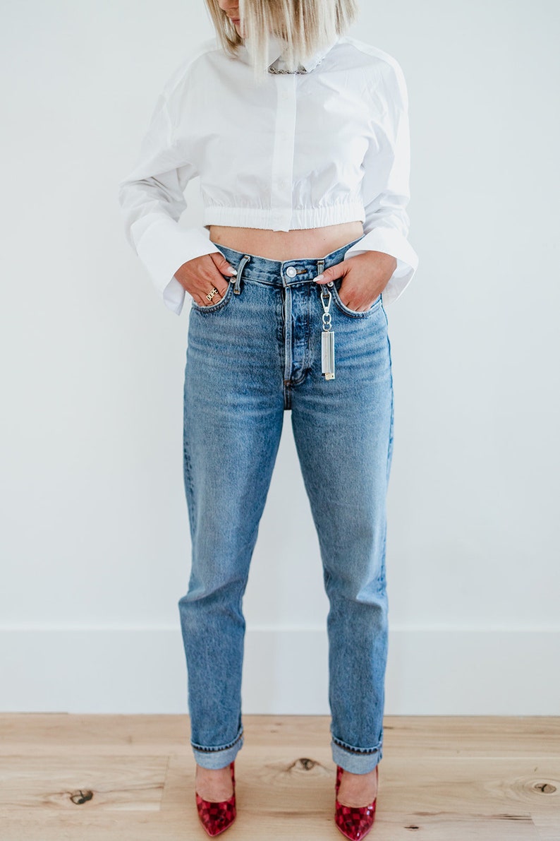May include: A woman wearing a white cropped button-down shirt with long sleeves, blue denim jeans, and red shoes. She is standing with her hands in her pockets.