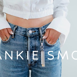 May include: A person wearing a white cropped shirt and blue jeans with a silver keychain attached to the right pocket. The keychain has a silver cylinder with the text "FRANKIE IS SMOKE" printed on it.