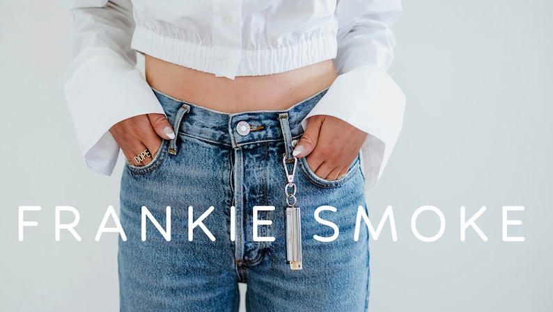 May include: A person wearing a white cropped shirt and blue jeans with a silver keychain attached to the right pocket. The keychain has a silver cylinder with the text "FRANKIE IS SMOKE" printed on it.