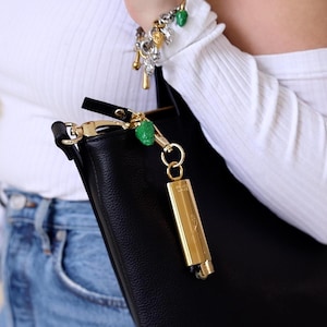 May include: A black leather tote bag with a gold zip and a gold key chain with a green charm. The bag is being held by a person wearing a white shirt and blue jeans.