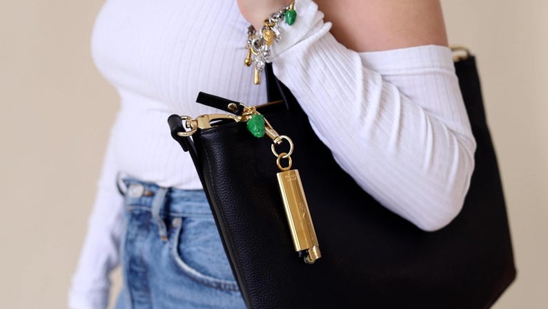 May include: A black leather tote bag with a gold zip and a gold key chain with a green charm. The bag is being held by a person wearing a white shirt and blue jeans.