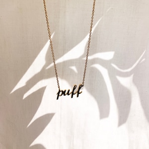 Op de afbeelding: Een gouden ketting met een hanger waarop in cursief schrift "puff" staat.