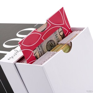 Puede incluir: Una caja blanca con un sobre de patr&oacute;n rojo y blanco que sobresale. La caja est&aacute; etiquetada "100 sobres". Un billete de d&oacute;lar es visible dentro del sobre.