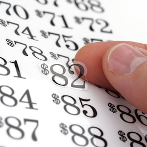 Puede incluir: Primer plano de una mano sosteniendo una pegatina con el n&uacute;mero 82. La pegatina se est&aacute; colocando en una hoja de papel con los n&uacute;meros 68, 70, 71, 72, 73, 74, 75, 77, 78, 81, 84, 85, 86, 87, 88 y 89 impresos en ella.