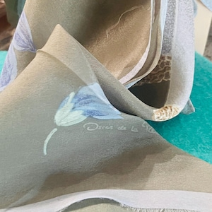 May include: A taupe-colored scarf featuring a light blue floral print and the text Oscar de la Renta printed in white. The fabric has a smooth texture with visible folds and a light-colored border along the edge of the material.