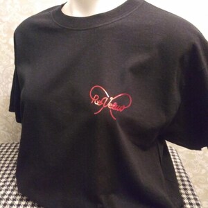 May include: A black short-sleeved t-shirt featuring a red metallic graphic on the chest. The graphic consists of a stylized infinity symbol shape with the text "ReVeluv" written in a cursive script font integrated into the center of the design.
