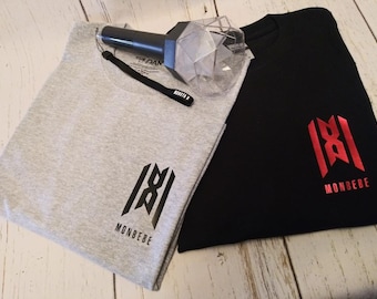 May include: Two t-shirts, one gray and one black, each with a logo that says "MONBEBE". A clear, geometric-shaped light stick with a black wrist strap is also present. The shirts are folded on a light-colored wooden surface.