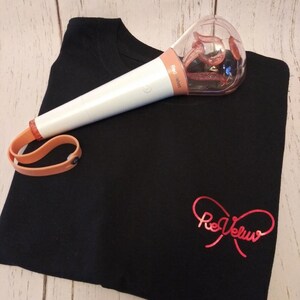 May include: A black t-shirt featuring a red metallic script logo that reads ReVeluv, paired with a white and peach-colored light stick. The light stick has a clear, rounded top containing a glittery, swirling interior element and a peach wrist strap.