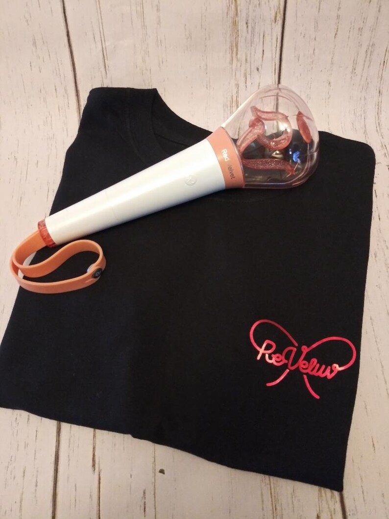 May include: A black t-shirt featuring a red metallic script logo that reads ReVeluv, paired with a white and peach-colored light stick. The light stick has a clear, rounded top containing a glittery, swirling interior element and a peach wrist strap.