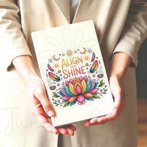 K&ouml;nnte beinhalten: Ein cremefarbenes Notizbuch mit den Worten "ALIGN and SHINE" in goldener Schrift. Das Cover zeigt eine farbenfrohe Lotusblume, Sonnen, Monde und Federn. Das Notizbuch wird von einer Person in einem beigen Blazer gehalten.