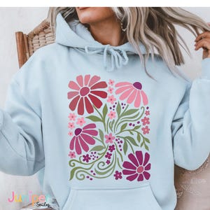 May include: Light blue hoodie featuring a floral design in shades of pink, red, and green. The graphic includes stylized flowers, leaves, and decorative swirls. The hoodie has a drawstring hood and a front pocket.