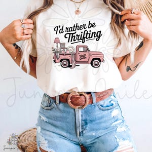 May include: A cream-coloured t-shirt with a pink vintage truck graphic, laden with thrifted items. The text "I'd rather be Thrifting" is above the truck, and "Thrift Queen" is on the door. The shirt is worn with denim shorts and a brown belt.