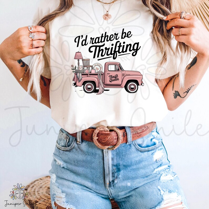 May include: A cream-coloured t-shirt with a pink vintage truck graphic, laden with thrifted items. The text "I'd rather be Thrifting" is above the truck, and "Thrift Queen" is on the door. The shirt is worn with denim shorts and a brown belt.