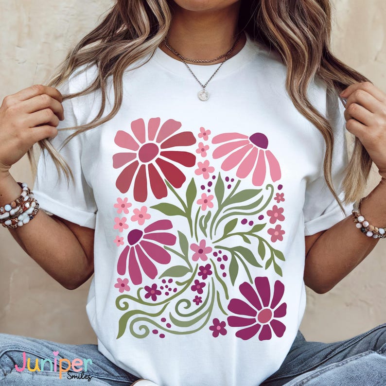 May include: White t-shirt featuring a floral design in shades of pink and red, with green leaves and stems. The design includes large stylized flowers and smaller blossoms. The text "Juniper Smiles" is at the bottom left.
