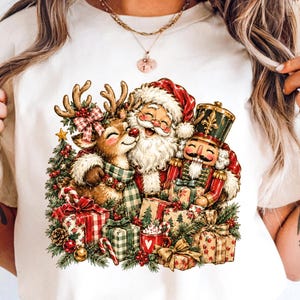 May include: A white t-shirt featuring a colourful graphic illustration of Santa Claus, a reindeer with a plaid scarf, and a nutcracker surrounded by wrapped Christmas gifts, pine branches, a small decorated tree, and a red mug with a heart.