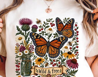 May include: A cream-colored t-shirt featuring a graphic print of two orange monarch butterflies, a mason jar filled with colorful wildflowers including purple thistles and daisies, small bees, and a banner with the text Wild & free.