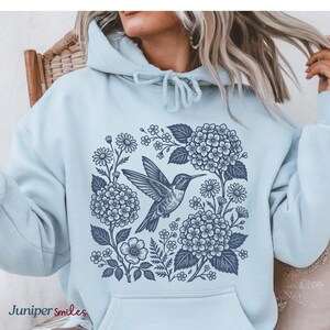 May include: A light blue hoodie featuring a navy blue hummingbird design surrounded by flowers and foliage. The text "Juniper smiles" is printed at the bottom left. The hoodie has a front pocket and a drawstring hood.