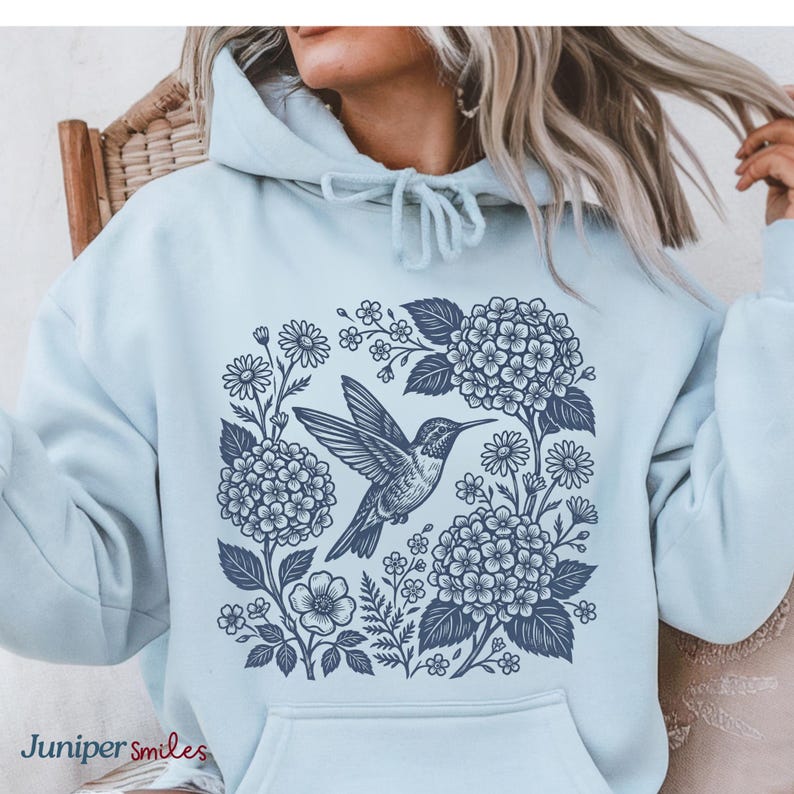 May include: A light blue hoodie featuring a navy blue hummingbird design surrounded by flowers and foliage. The text "Juniper smiles" is printed at the bottom left. The hoodie has a front pocket and a drawstring hood.