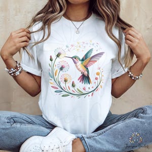 Op de afbeelding: Wit T-shirt met een kleurrijk kolibrie-ontwerp. De illustratie toont een kolibrie met levendige vleugels, omringd door bloemen en bladeren. Het ontwerp is gecentreerd op het shirt, waardoor een visueel aantrekkelijke afbeelding ontstaat.
