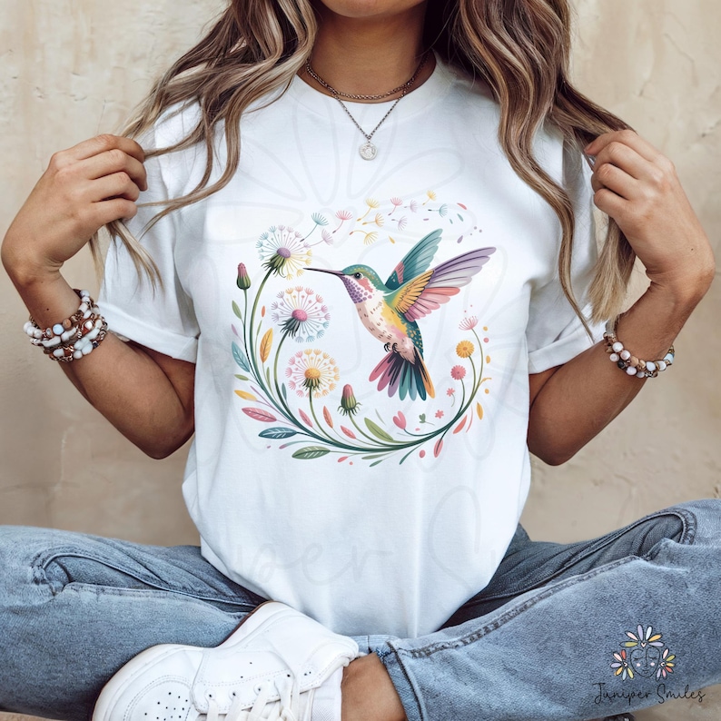 Op de afbeelding: Wit T-shirt met een kleurrijk kolibrie-ontwerp. De illustratie toont een kolibrie met levendige vleugels, omringd door bloemen en bladeren. Het ontwerp is gecentreerd op het shirt, waardoor een visueel aantrekkelijke afbeelding ontstaat.