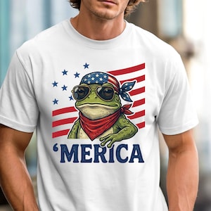 Könnte beinhalten: Weißes T-Shirt mit einer Cartoon-Frösche, die eine Sonnenbrille, ein rotes Bandana und ein Stirnband mit einem amerikanischen Flaggenmuster trägt. Der Frosch steht vor einem rot-weiß gestreiften Hintergrund mit blauen Sternen. Das Wort 'Merica ist darunter gedruckt.