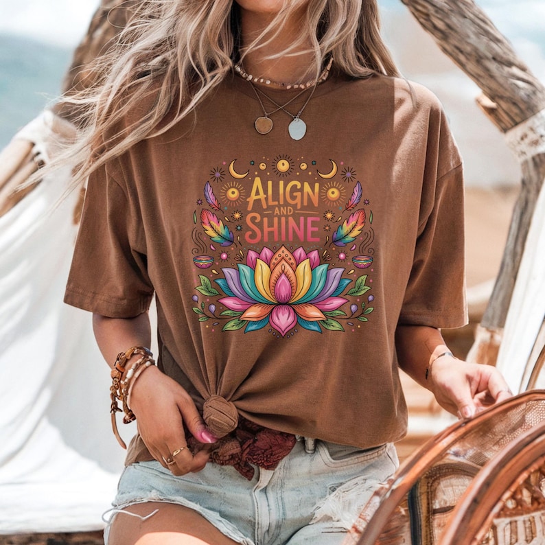 K&ouml;nnte beinhalten: Braunes T-Shirt mit dem Text "Align and Shine" &uuml;ber einem farbenfrohen Lotusblumen-Design. Das Shirt hat kurze &Auml;rmel und eine geknotete Vorderseite. Das Design beinhaltet Regenbogen, Monde und Sonnen.