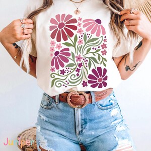 May include: Cream-colored t-shirt featuring a floral design in shades of pink and purple, with green leaves and stems. The design includes large and small flowers, and the text "Juniper Smiles" is visible at the bottom.