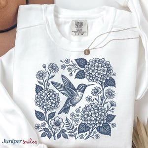 May include: White sweatshirt with a blue floral and hummingbird design. The graphic features a hummingbird surrounded by flowers and foliage. A gold necklace with a small pendant is also visible. The brand name "Juniper smiles" is printed on the bottom left.