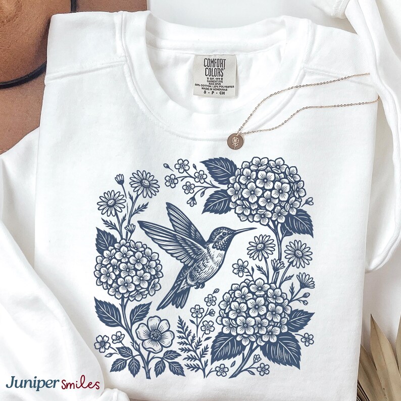 May include: White sweatshirt with a blue floral and hummingbird design. The graphic features a hummingbird surrounded by flowers and foliage. A gold necklace with a small pendant is also visible. The brand name "Juniper smiles" is printed on the bottom left.