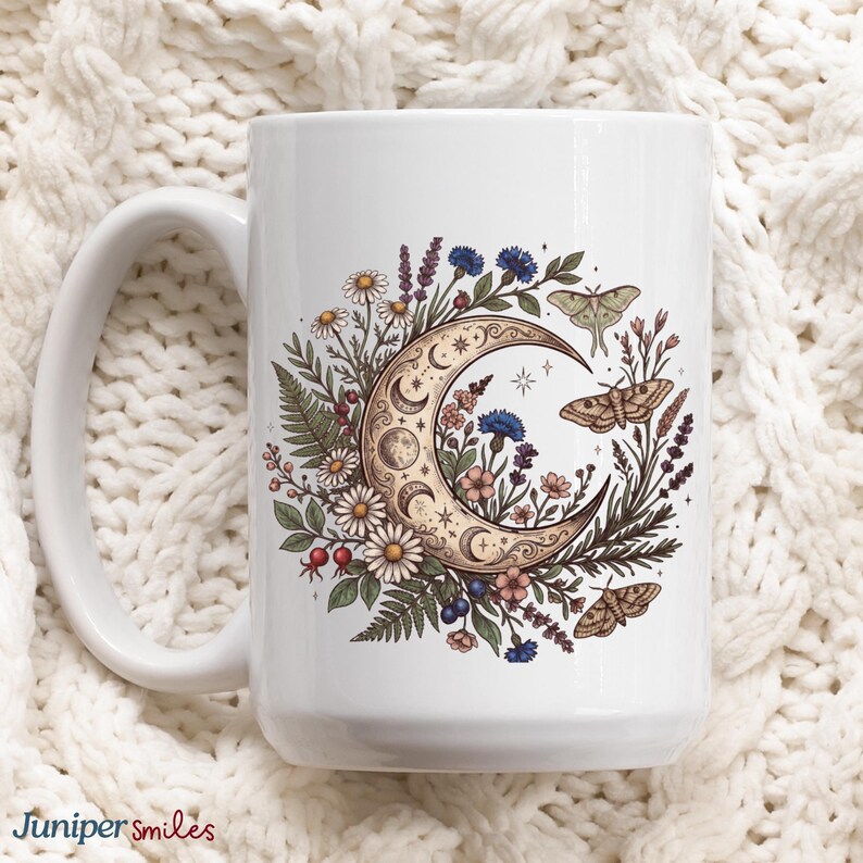 May include: White ceramic mug featuring a crescent moon design filled with flowers, leaves, and moths. The design includes daisies, blue cornflowers, and other floral elements. The mug has a large handle and the text "Juniper smiles" at the bottom.