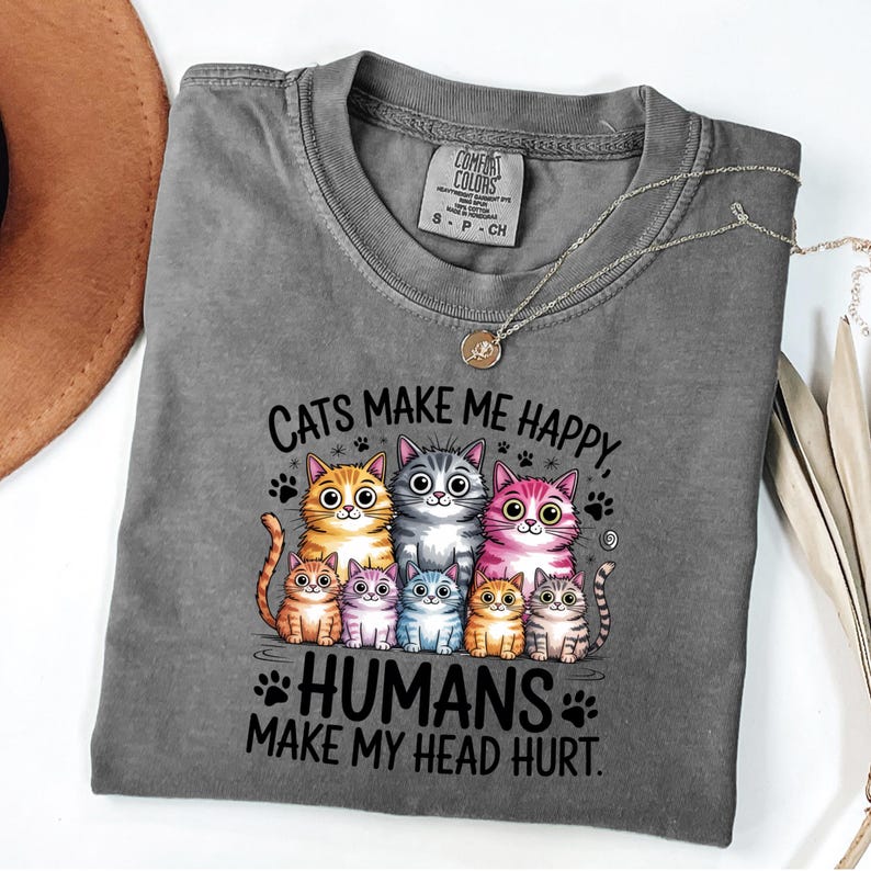 Puede incluir: Una camiseta gris Comfort Colors con un gr&aacute;fico de siete gatos de dibujos animados en varios colores. El texto dice "Cats make me happy, humans make my head hurt." Un collar dorado est&aacute; drapeado sobre la camisa.