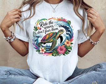 Pode incluir: Camiseta branca com um design colorido de uma tartaruga marinha nadando em frente a uma onda. O design inclui o texto "Ride the waves, don't fear them". Flores e folhagens tropicais cercam a tartaruga e a onda.