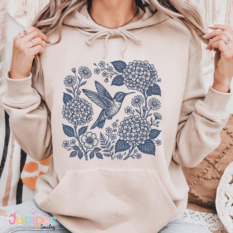 May include: Beige hoodie with a blue floral and hummingbird design. The graphic features a hummingbird in flight surrounded by flowers and leaves. The hoodie has a drawstring hood and a front pocket.