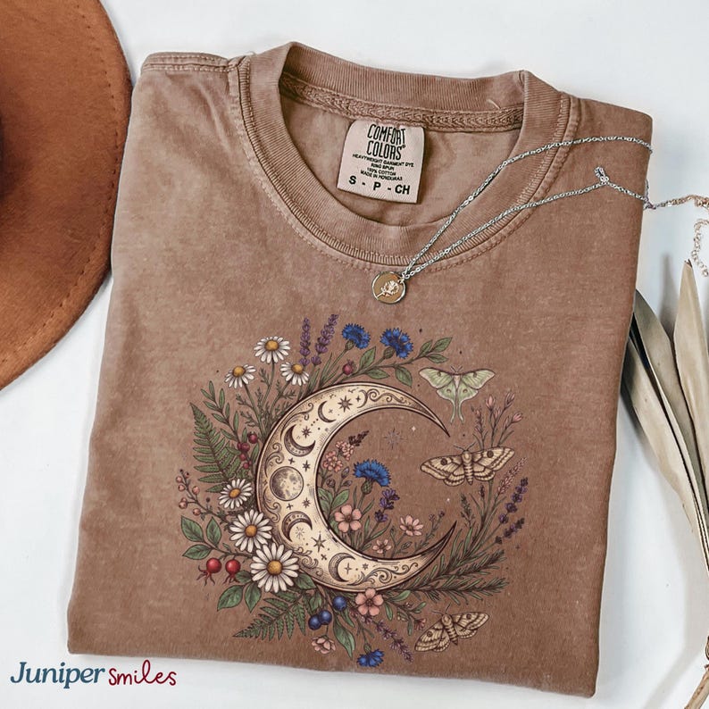 May include: A brown Comfort Colors t-shirt with a crescent moon and floral design. The graphic includes daisies, blue flowers, and moths. A silver necklace with a small pendant rests on the shirt. The shirt is folded and displayed on a white surface.