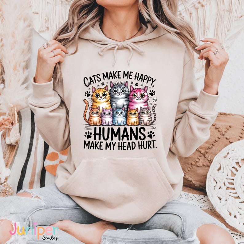 Puede incluir: Sudadera con capucha beige con un dise&ntilde;o de gatos de dibujos animados en varios colores. El texto "CATS MAKE ME HAPPY, HUMANS MAKE MY HEAD HURT" est&aacute; impreso debajo de los gatos.