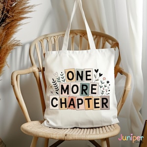 May include: A cream-coloured tote bag featuring the words "ONE MORE CHAPTER" in colourful block letters, with floral and heart motifs. The bag rests on a light brown wicker chair.