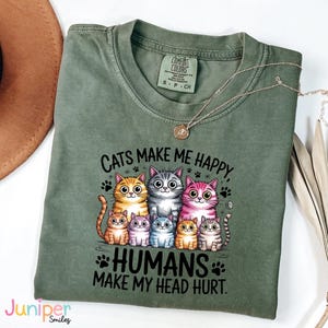 Puede incluir: Una camiseta verde salvia con el texto "Cats make me happy, humans make my head hurt". El gr&aacute;fico presenta un grupo de gatos de dibujos animados en varios colores. Un collar dorado est&aacute; drapeado sobre la camiseta.