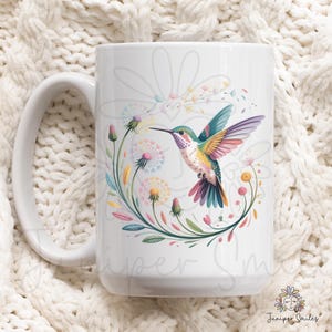Op de afbeelding: Witte keramische mok met een kleurrijk ontwerp van een kolibrie in vlucht, omringd door bloemen en paardenbloemmotieven. De vogel heeft groene, gele en roze veren. De mok heeft een groot handvat en is geschikt voor warme dranken.